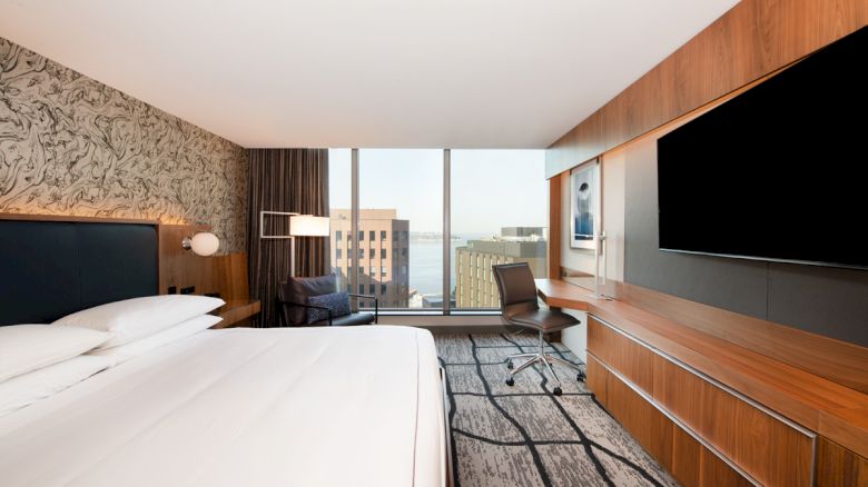 A modern hotel room with a large bed, wall-mounted TV, desk, chair, and a view of city buildings through a large window is shown.