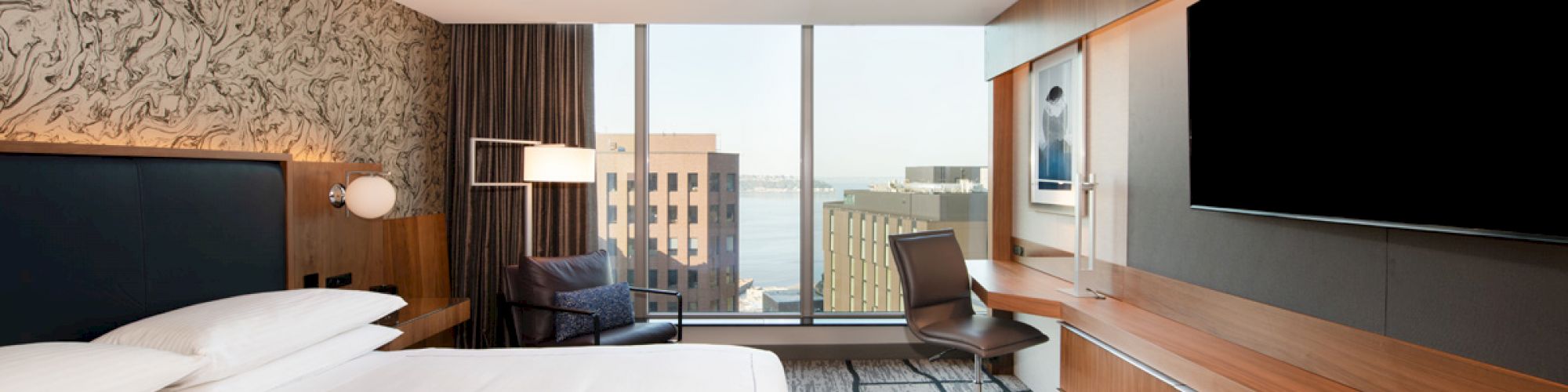 The image shows a modern hotel room with a large bed, flat-screen TV, desk, chair, and a window offering a view of city buildings outside.