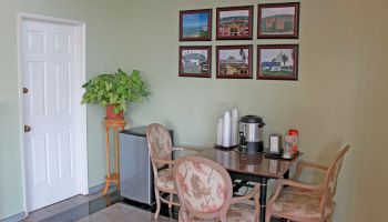 A small seating area with a table, four chairs, coffee station, framed pictures on the wall, a mini-fridge, a potted plant, and a closed door.