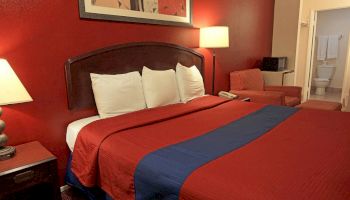 A hotel room with a bed, nightstands, lamps, wall art, an armchair, a small table, and a view into a bathroom at the back.