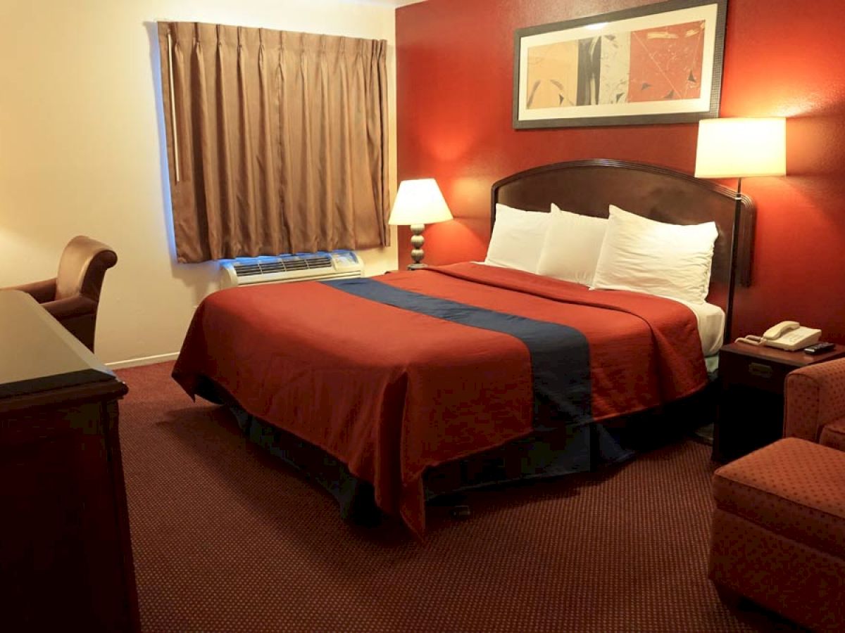 A hotel room with a king-sized bed, two nightstands, a chair, a window with curtains, a TV, and a table lamp, all in warm earth tones.