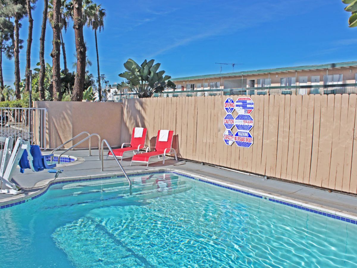 The image shows an outdoor swimming pool area with two red loungers, a wooden fence with signs, and palm trees in the background.