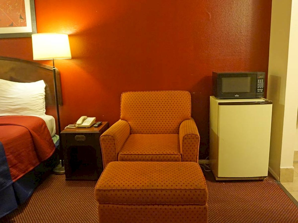 The image shows a hotel room with a bed, nightstand, lamp, armchair, ottoman, mini-fridge, microwave, and an area for hanging clothes.