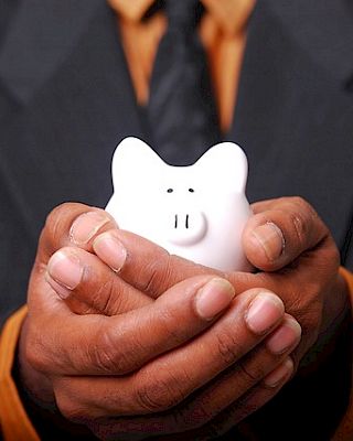 A person in a suit holds a small white piggy bank with both hands against a dark background, symbolizing saving or financial security.