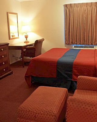 The image shows a hotel room with a bed, TV on a dresser, desk with chair, armchair, ottoman, and lamps, all in warm, earthy tones.