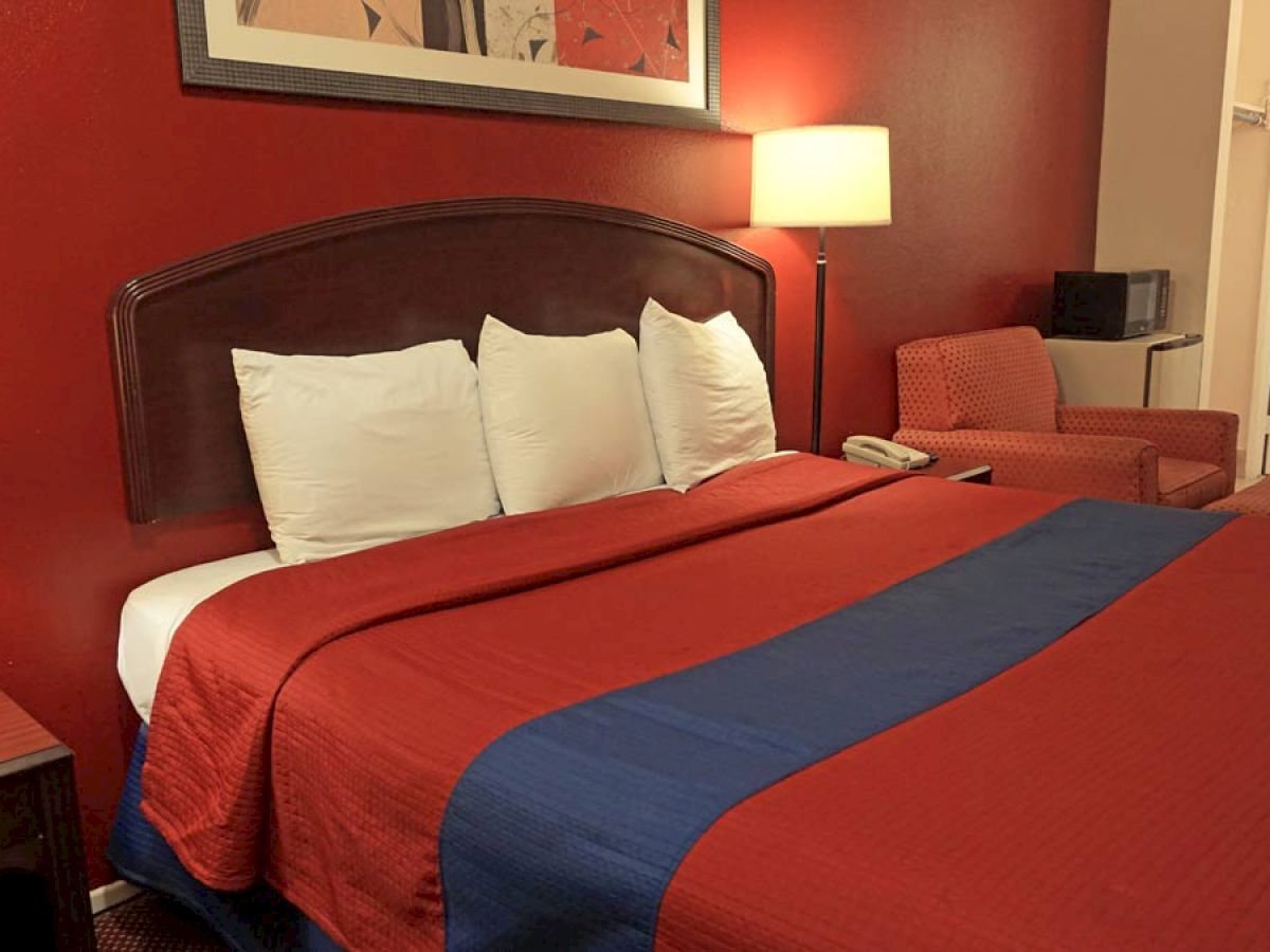 The image shows a hotel room with a neatly made bed, bedside lamps, a red accent wall, an armchair, and an attached bathroom.